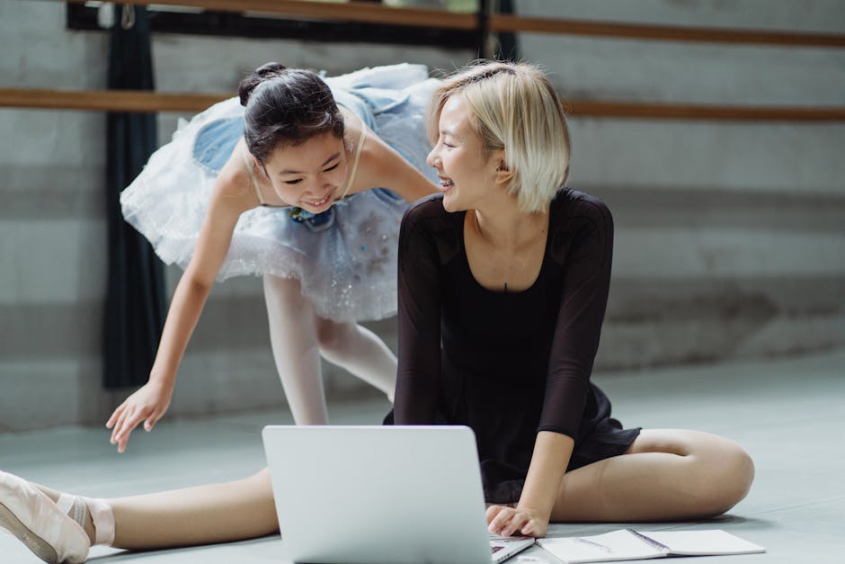 Cheerful Asian ballerina in tutu skirt with personal trainer searching information in netbook while laughing together in ballet studio